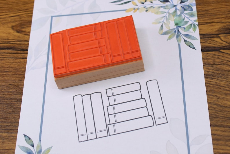 K&ouml;nnte beinhalten: Ein orangefarbener Gummistempel mit Holzsockel, der ein B&uuml;cherstapel-Design zeigt. Der Stempel liegt auf wei&szlig;em Papier mit einer gedruckten Umrisszeichnung von B&uuml;chern. Der Hintergrund ist eine Holzoberfl&auml;che.