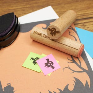 Op de afbeelding: Twee cilindrische houten stempels met de tekst SCRAWL STUDIO en een spookicoon gegraveerd op de uiteinden. De ene stempel ligt horizontaal, terwijl de andere er diagonaal bovenop rust. Twee kleine vierkante papiertjes tonen zwarte spookafdrukken.