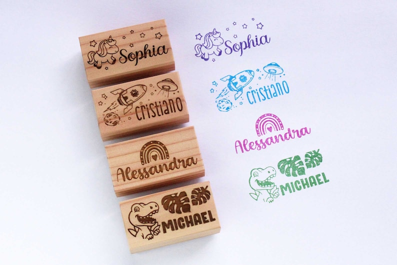 May include: Wooden stamps with engraved designs and names. The stamps feature a unicorn, rocket, rainbow, and dinosaur, each paired with a name: Sophia, Cristiano, Alessandra, and Michael. The stamped impressions are in various colours.