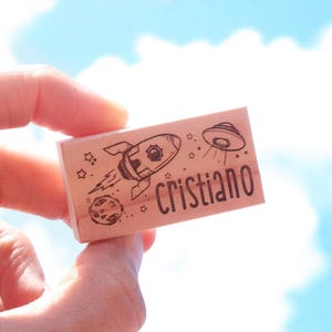 May include: A wooden stamp with a rocket, planet, and UFO design, plus the name "Cristiano". The stamp is held up against a blue sky with clouds. The stamp is a fun, space-themed item.