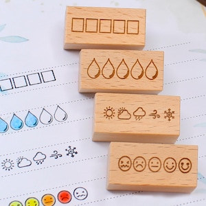 K&ouml;nnte beinhalten: Vier Holzstempel mit eingravierten Symbolen zur Verfolgung von Stimmung, Wetter und anderen Metriken. Die Stempel umfassen K&auml;stchen, Wassertropfen, Sonne, Wolke, Wind, Schneeflocke und Gesichter. Die Stempel befinden sich auf einem Papier mit floralem Muster.