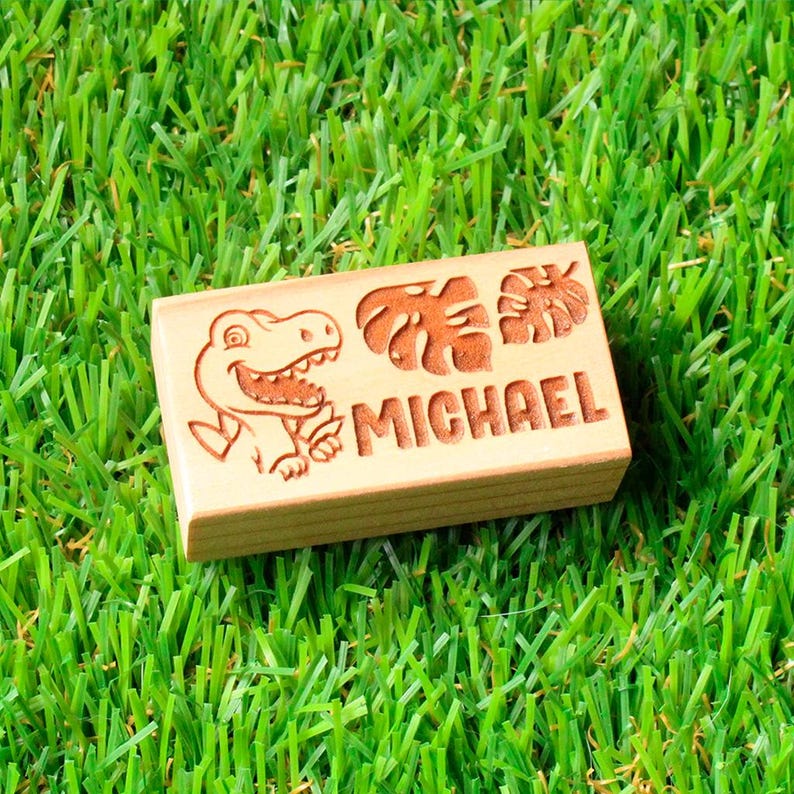 May include: A rectangular wooden block engraved with the name "MICHAEL". The block features a cartoon dinosaur and leaf designs. The block is set against a backdrop of green artificial grass.