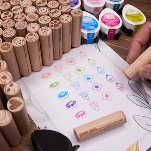 Può includere: Una collezione di timbri cilindrici in legno con varie icone incise sulle estremità, tra cui cibo, animali e forme. Sono presenti diversi cuscinetti di inchiostro colorati e un foglio di carta bianco mostra le impronte di questi disegni.