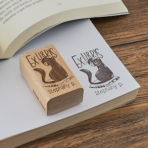 May include: A wooden "Ex Libris" stamp with a cat illustration and the name "Stephany p." is next to a stamped impression on a book page. An open book rests in the background on a wooden surface.