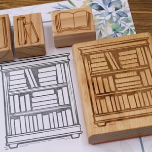 K&ouml;nnte beinhalten: Holzstempel mit Buch- und B&uuml;cherregal-Designs. Die Stempel sind aus hellem Holz gefertigt und zeigen detaillierte Illustrationen von B&uuml;chern und einem B&uuml;cherregal. Eine Zeichnung eines B&uuml;cherregals ist ebenfalls zu sehen.