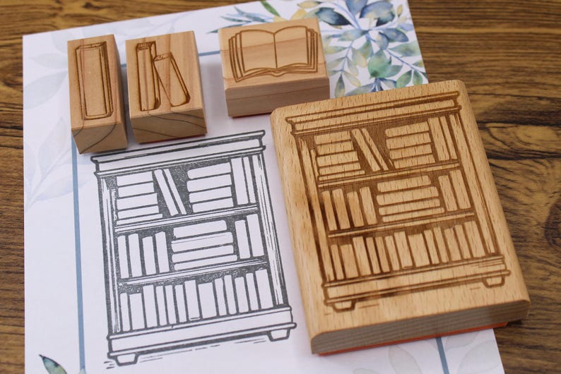 K&ouml;nnte beinhalten: Holzstempel mit Buch- und B&uuml;cherregal-Designs. Die Stempel sind aus hellem Holz gefertigt und zeigen detaillierte Illustrationen von B&uuml;chern und einem B&uuml;cherregal. Eine Zeichnung eines B&uuml;cherregals ist ebenfalls zu sehen.