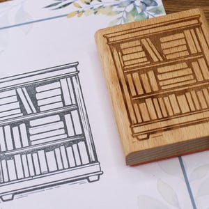 K&ouml;nnte beinhalten: Ein Holzstempel mit einem B&uuml;cherregal-Design neben einem gedruckten Bild desselben B&uuml;cherregals. Der Stempel ist rechteckig mit einer detaillierten B&uuml;cherregal-Gravur. Das gedruckte Bild ist eine Schwarz-Wei&szlig;-Strichzeichnung.
