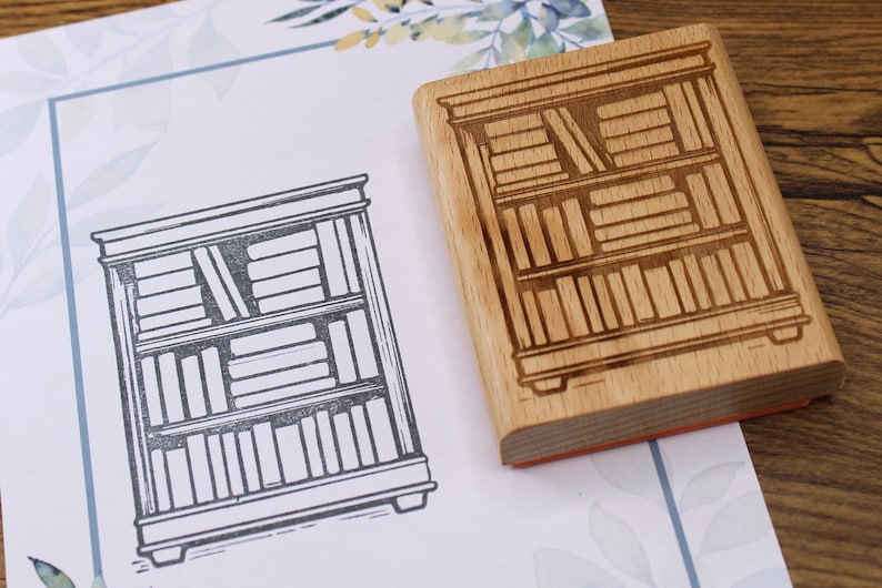 K&ouml;nnte beinhalten: Ein Holzstempel mit einem B&uuml;cherregal-Design neben einem gedruckten Bild desselben B&uuml;cherregals. Der Stempel ist rechteckig mit einer detaillierten B&uuml;cherregal-Gravur. Das gedruckte Bild ist eine Schwarz-Wei&szlig;-Strichzeichnung.