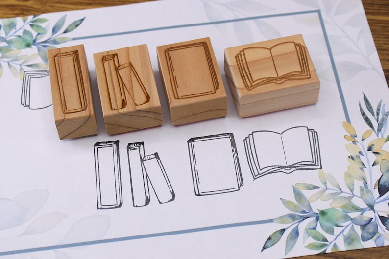 K&ouml;nnte beinhalten: Holzstempel mit Buchmotiven. Die Stempel sind rechteckig mit ge&auml;tzten Buchillustrationen. Unter den Stempeln befinden sich passende Buchumrisse. Der Hintergrund ist ein wei&szlig;es Papier mit floralem Design.