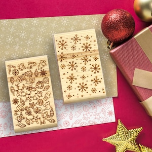 Könnte beinhalten: Zwei rechteckige Holzstempel mit lasergravierten Weihnachtsmotiven. Ein Stempel zeigt eine Ansammlung verschiedener Weihnachtssymbole wie Rentiere, Ornamente und Geschenke, während der andere ein wiederkehrendes Schneeflockenmuster aufweist.