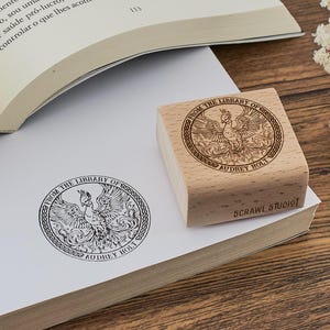 Puede incluir: Un sello de goma de madera con un diseño circular que presenta un fénix con el texto "From the Library of Audrey Holt" y "Scrawl Studio".