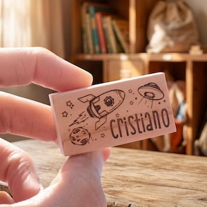 May include: A wooden stamp with a rocket, planet, and UFO design, plus the name "Cristiano". The stamp is held in a hand. The background shows a wooden shelf with books. The stamp is approximately 7.5 cm long.