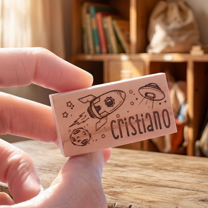May include: A wooden stamp with a rocket, planet, and UFO design, plus the name "Cristiano". The stamp is held in a hand. The background shows a wooden shelf with books. The stamp is approximately 7.5 cm long.