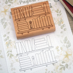 K&ouml;nnte beinhalten: Ein Holzstempel mit einem B&uuml;cherregal-Design, einschlie&szlig;lich gestapelter B&uuml;cher und einer Kerze. Der Stempel liegt auf floral gemustertem Papier mit einer passenden B&uuml;cherregal-Kontur. B&uuml;cher sind im Hintergrund zu sehen.