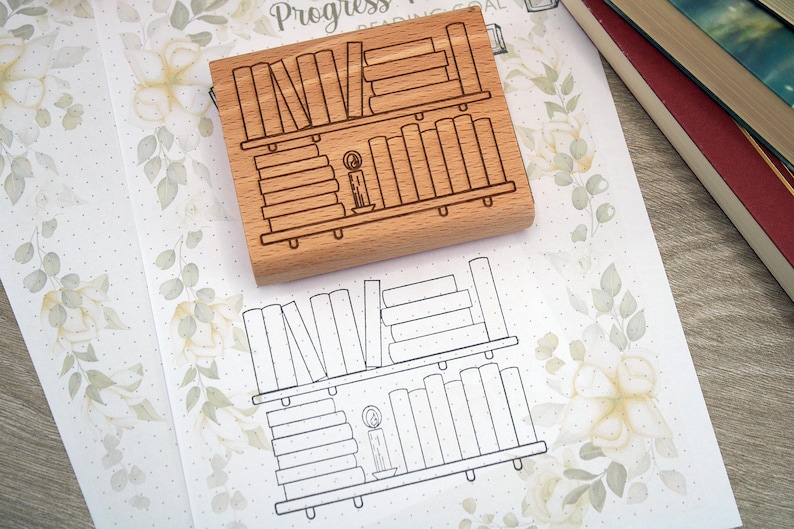 K&ouml;nnte beinhalten: Ein Holzstempel mit einem B&uuml;cherregal-Design, einschlie&szlig;lich gestapelter B&uuml;cher und einer Kerze. Der Stempel liegt auf floral gemustertem Papier mit einer passenden B&uuml;cherregal-Kontur. B&uuml;cher sind im Hintergrund zu sehen.