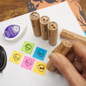 Op de afbeelding: Vier cilindervormige houten stempels met schedelontwerpen, elk gemarkeerd met de tekst 5CRAWL STUDIO. Een paars Memento Grape Jelly inktkussen en vijf kleine vierkante papieren briefjes in groen, blauw, geel en roze met zwarte schedelafdrukken.