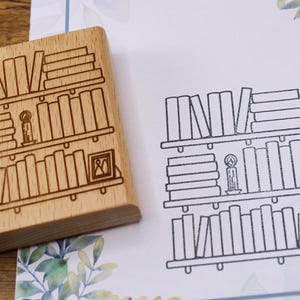 K&ouml;nnte beinhalten: Ein Holzstempel mit einem B&uuml;cherregal-Design, neben einem gestempelten Bild desselben Designs. Der Stempel zeigt drei B&uuml;cherregale, eine Kerze und ein gerahmtes Bild. Das gestempelte Bild ist in schwarzer Tinte auf wei&szlig;em Papier.