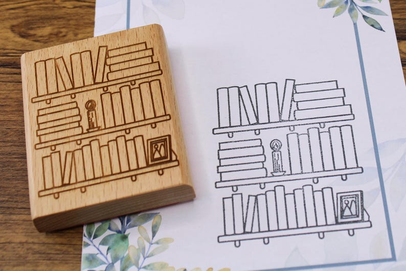 K&ouml;nnte beinhalten: Ein Holzstempel mit einem B&uuml;cherregal-Design, neben einem gestempelten Bild desselben Designs. Der Stempel zeigt drei B&uuml;cherregale, eine Kerze und ein gerahmtes Bild. Das gestempelte Bild ist in schwarzer Tinte auf wei&szlig;em Papier.