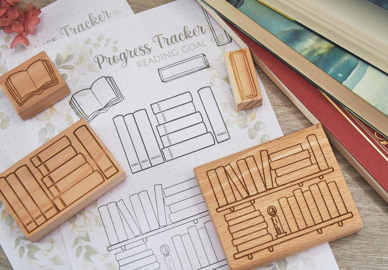 K&ouml;nnte beinhalten: Holzstempel mit Buch- und B&uuml;cherregal-Designs werden auf einem Papier mit floralem Muster mit dem Text "Progress Tracker Reading Goal" gezeigt. Im Hintergrund sind auch mehrere B&uuml;cher zu sehen.