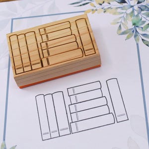 K&ouml;nnte beinhalten: Ein Holzstempel mit rechteckiger Form, der ein Design aus mehreren gestapelten B&uuml;chern zeigt. Der Stempel liegt auf einem wei&szlig;en Papier mit einer gedruckten Umrisszeichnung von B&uuml;chern. Das Papier hat einen blauen Rand und florale Akzente.