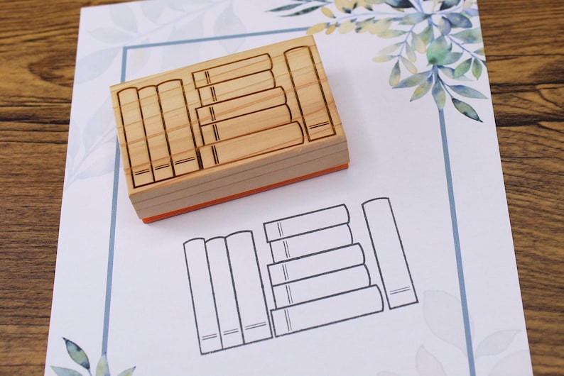 K&ouml;nnte beinhalten: Ein Holzstempel mit rechteckiger Form, der ein Design aus mehreren gestapelten B&uuml;chern zeigt. Der Stempel liegt auf einem wei&szlig;en Papier mit einer gedruckten Umrisszeichnung von B&uuml;chern. Das Papier hat einen blauen Rand und florale Akzente.