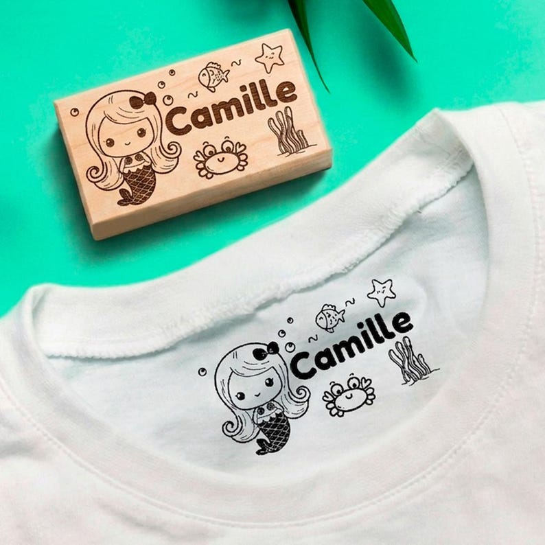 May include: A wooden stamp featuring a mermaid design and the name "Camille" is displayed beside a white t-shirt with the same design printed on it. The stamp includes a mermaid, fish, crab, bubbles, and seaweed.