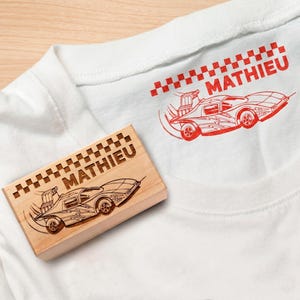May include: White t-shirt featuring a red race car design and the name "MATHIEU". A wooden stamp with the same car and name is next to the shirt.
