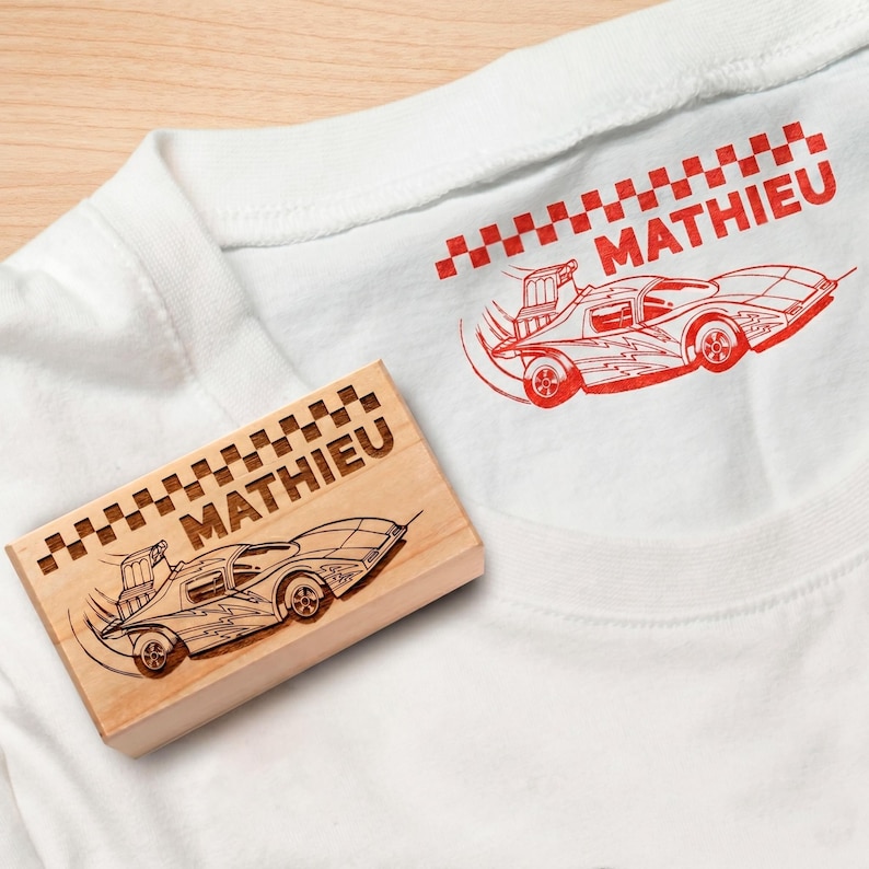 May include: White t-shirt featuring a red race car design and the name "MATHIEU". A wooden stamp with the same car and name is next to the shirt.