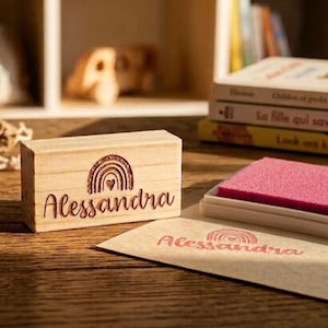 May include: A wooden stamp featuring the name "Alessandra" in a script font, with a rainbow and heart design. A pink ink pad and a stamped image of the name and rainbow are also visible. The stamp is on a wooden surface.