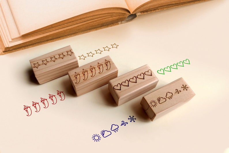 K&ouml;nnte beinhalten: Vier Holzstempel mit Stern-, Chili-, Herz- und Wettersymbol-Designs. Jeder Stempel hat eine entsprechende Reihe von gestempelten Bildern in roter, gr&uuml;ner und blauer Tinte. Ein offenes Buch befindet sich im Hintergrund.