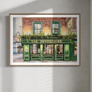 Op de afbeelding: Een ingelijste aquareltekening van de gevel van een pub, met de gouden tekst "THE DEVONSHIRE". Het gebouw heeft een groene buitenkant, bakstenen, groene kozijnen en decoratief groen. Het kunstwerk is gepresenteerd in een houten lijst.