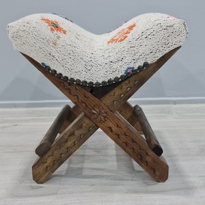 May include: A white and brown folding stool with a patterned cushion. The stool has a carved wooden frame and a white fabric cushion with colorful embroidery.
