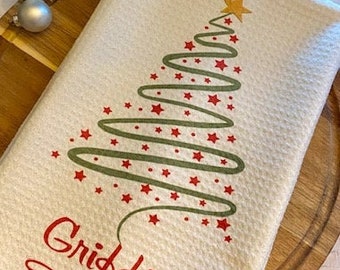May include: White waffle-weave kitchen towel with a Christmas tree design. The tree is made of a green swirled line with red star accents and a gold star on top. The words "Griffin Family" are in red script.