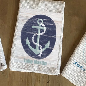May include: Three white kitchen towels with blue and gray nautical designs. The first towel says "Welcome to our Lake House" with an anchor. The second towel has a blue circle with a white anchor and rope and says "Lake Martin". The third towel says "Life is better at the Lake" with an anchor and says "Lake Martin".