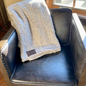 May include: A plush, white throw blanket with a brown leather patch that says "J.Crew Mercantile" is draped over a black leather armchair.