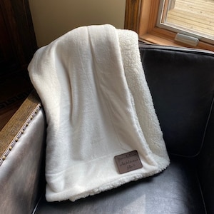 May include: White sherpa throw blanket with a brown leather tag that says "Handmade by [unclear text] for [unclear text]"