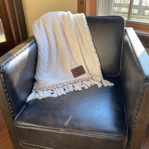 May include: A white faux fur throw blanket with a brown leather patch and fringe trim, draped over a black leather armchair with a wooden frame.