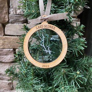 May include: A wooden Christmas ornament with a clear plastic insert featuring a map of Lake Oconee, Georgia. The ornament is inscribed with the text "LAKE OCONEE", "33.4303° N 83.1938° W", and "2022".