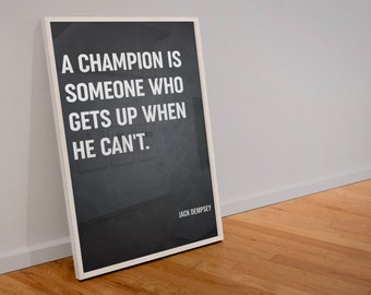May include: A framed poster with a black background and white text. The quote reads: "A CHAMPION IS SOMEONE WHO GETS UP WHEN HE CAN'T." The author's name, Jack Dempsey, is at the bottom. The frame is white.