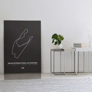 Puede incluir: Un p&oacute;ster negro con un contorno blanco del circuito del Aut&oacute;dromo Internacional de Miami. El p&oacute;ster incluye el nombre de la pista, detalles y una peque&ntilde;a bandera estadounidense. El p&oacute;ster se exhibe en una habitaci&oacute;n con mesas auxiliares, una l&aacute;mpara y una planta.