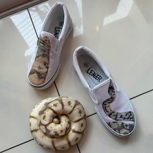 May include: White canvas slip-on shoes with a snake skin design on the top. The shoes are next to a coiled snake. The shoes have a black sole and the brand name "LEINIER" is printed on the insole.