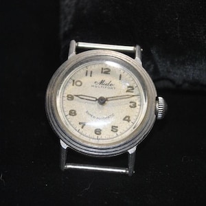 Pu&ograve; includere: Un orologio vintage Mido Multifort con cassa color argento e quadrante color crema. L'orologio presenta numeri neri, lancette e la scritta "SUPER-AUTOMATIC". Ha una corona a destra e attacchi per cinturino argentati.