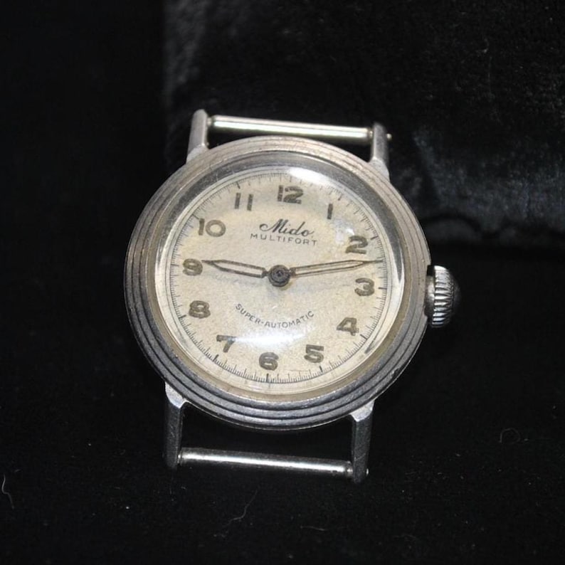 Pu&ograve; includere: Un orologio vintage Mido Multifort con cassa color argento e quadrante color crema. L'orologio presenta numeri neri, lancette e la scritta "SUPER-AUTOMATIC". Ha una corona a destra e attacchi per cinturino argentati.