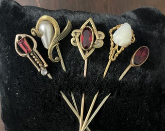 Può includere: Una collezione di antichi spilloni da cappello esposti su una superficie di velluto nero. Gli spilloni presentano vari design, tra cui una perla, pietre preziose rosse e decorazioni in metallo ornate. Alcuni spilloni hanno una finitura dorata, mentre altri sono argentati. È visibile anche un gruppo di spilloni da cappello semplici.