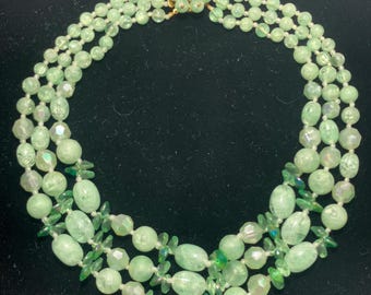 May include: A three-strand necklace featuring light green beads of various shapes and sizes. The necklace has a gold-colored clasp and is composed of round, oval, and faceted beads, with small green accents. The necklace is displayed against a black background.