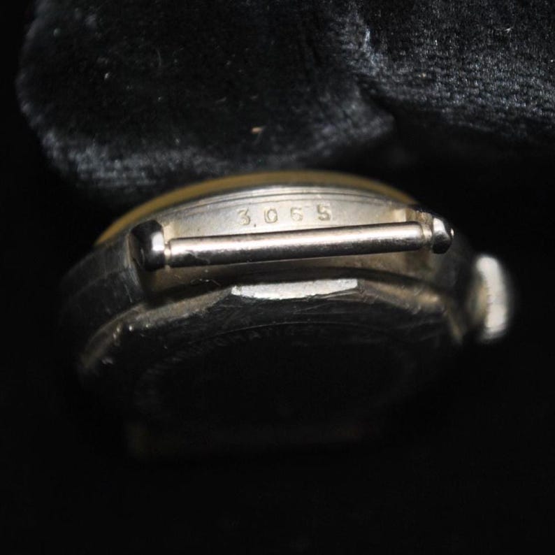 Pu&ograve; includere: Primo piano di un orologio da polso vintage con cassa color argento e quadrante ingiallito. Il numero "3065" &egrave; inciso sul cinturino dell'orologio. L'orologio &egrave; ambientato su uno sfondo scuro.