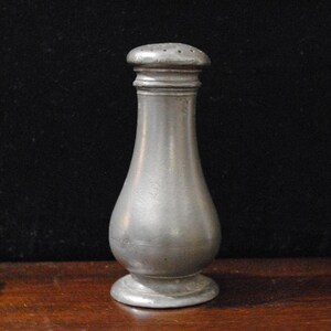 May include: A vintage, silver-colored salt shaker with a rounded body and a small, perforated lid. The shaker has a classic, hourglass shape, and sits on a small, circular base. The metal has a slightly aged appearance.