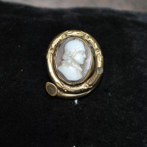 May include: A vintage cameo brooch featuring a white profile portrait set in an oval frame. The frame is gold-toned with an ornate, engraved design. The brooch has a curved shape with a small clasp.