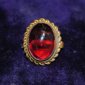 May include: A gold-toned ring with an oval, deep red gemstone. The gemstone is set in a gold frame with a twisted rope design around the edge. The ring is on a dark purple surface.