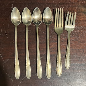 May include: A set of five silver-colored spoons and two forks. The spoons have long, slender handles and oval bowls. The forks have four tines. The silverware is arranged on a dark wooden surface.